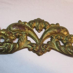 May include: Ornate, antique-style metal furniture hardware in a gold and bronze finish. The decorative piece features a symmetrical design with scrolling details and two circular openings. The finish shows signs of wear.