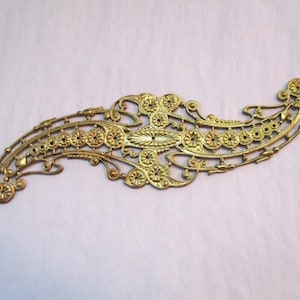 May include: A gold-toned metal filigree design with a central oval shape and intricate floral patterns. The design is reminiscent of a vintage or antique style.