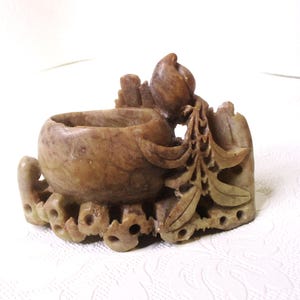 Può includere: Un piccolo bruciatore di incenso in pietra finemente scolpito con un top a forma di ciotola e un motivo floreale. Il bruciatore è fatto di pietra marrone chiaro con una texture ruvida.