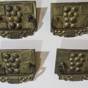 以下が含まれることがあります： 正方形のベースと円と正方形の装飾的なデザインが施された、アンティークな真鍮製のキャビネットプルが4つ。プルにはループハンドルが付いています。