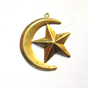 May include: A gold-colored pendant featuring a crescent moon and a five-pointed star. The star is centered within the moon's curve. The pendant has a small loop at the top for hanging. The metal has a slightly aged appearance.