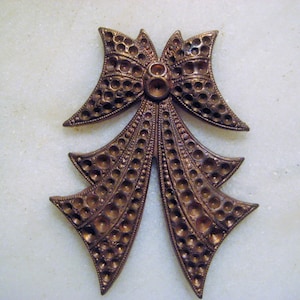 May include: A bronze-colored bow-shaped brooch with a detailed openwork design. The brooch features a central knot with circular accents and cascading ribbon-like elements, all with a textured, aged appearance. The bow is symmetrical and appears to be made of metal.