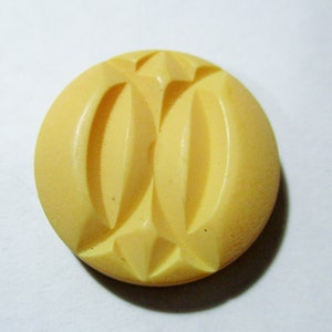 May include: A yellow plastic button with a decorative design. The button has a raised, textured pattern that resembles a stylized flower or leaf.