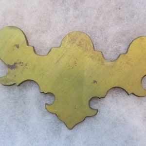 Könnte beinhalten: Ein goldfarbener, dekorativer Metallbesatz mit einem gewellten Rand und einem dreispitzigen Design. Der Besatz hat ein abgenutztes, gealtertes Aussehen.