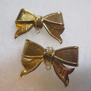 May include: Two gold-tone bow-shaped metal embellishments with a textured finish. Each bow has a small loop at the bottom.