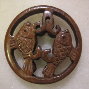 以下が含まれることがあります： 3匹の魚が彫刻された丸い木製のペンダント。魚は精巧に細工されており、茶色です。