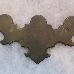 Könnte beinhalten: Ein dekoratives Messing-Hardware-Teil mit einem gewellten Rand und zwei Löchern. Das Teil hat die Form einer stilisierten Fleur-de-lis.