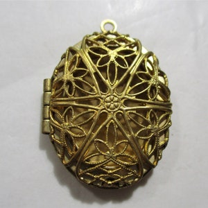 Puede incluir: Un medallón ovalado dorado con un delicado diseño de filigrana. El medallón tiene una abertura con bisagra y un bucle en la parte superior para sujetar una cadena.