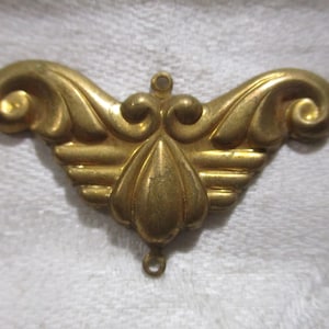 May include: A gold-colored metal ornament with a decorative, ornate design. The ornament has a teardrop shape with two loops at the top and bottom.