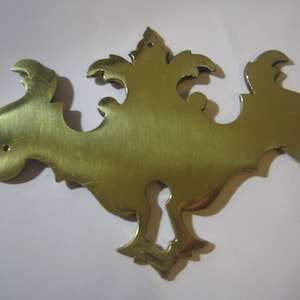 Op de afbeelding: Een gouden metalen sleutelgatbedekking met een decoratief, sierlijk ontwerp. De bedekking heeft een sleutelgat in het midden en een symmetrisch, ingewikkeld patroon.