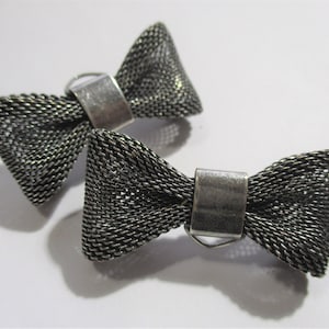 Könnte beinhalten: Zwei silberfarbene Accessoires in Form einer Fliege. Die Schleifen bestehen aus einem gewebten, netzartigen Material. Jede Schleife hat einen rechteckigen Metallverschluss in der Mitte. Die Accessoires werden auf einer weißen Oberfläche präsentiert.