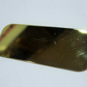 May include: A rectangular, gold-colored metal object with rounded corners. The surface has minor scratches and imperfections, reflecting light. The item appears to be a small, decorative piece or component.