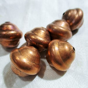 Puede incluir: Un grupo de seis cuentas de color cobre con un diseño de espiral. Las cuentas están dispuestas en semicírculo sobre una superficie blanca.