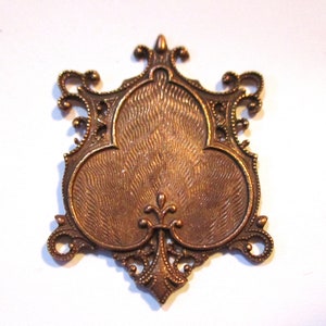 May include: A bronze-colored metal charm in the shape of a stylized clover with intricate filigree details. The charm has a textured surface and a decorative design.