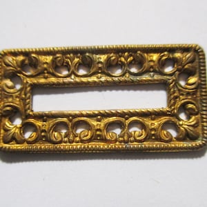 May include: A gold-colored metal rectangular frame with an ornate design. The frame has a rectangular opening in the center.