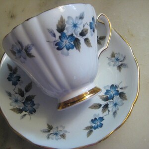 May include: A white teacup and saucer set with a blue and gray floral pattern. The cup has a gold rim and the saucer has a gold edge.