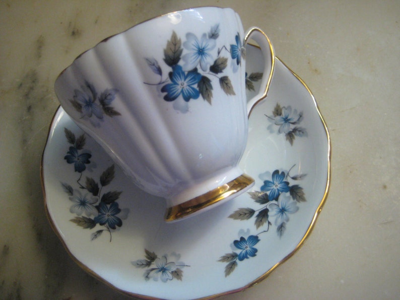 May include: A white teacup and saucer set with a blue and gray floral pattern. The cup has a gold rim and the saucer has a gold edge.