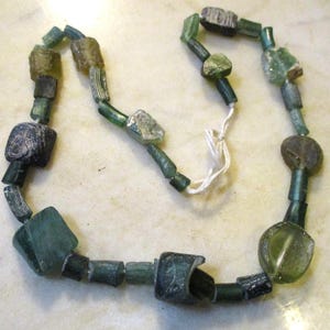 May include: A necklace composed of various shaped beads in shades of green, blue, and brown. The beads are strung together with a white string. The beads have a weathered, antique appearance.
