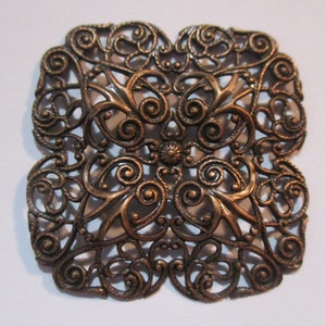 May include: Ornate, square-shaped bronze metal filigree. The openwork design features swirling patterns, floral accents, and a central flower. The edges have a rope-like texture. This decorative piece could be used for jewelry or other craft projects.