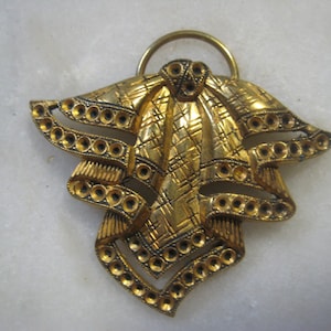 May include: A gold-toned metal pendant with a decorative design of a bow with a textured surface. The pendant has a loop at the top for hanging.