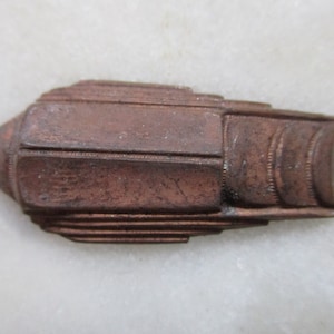 May include: A small, copper-colored metal object with a geometric design. It has a pointed end and a rounded edge. The object is about 2 inches long.