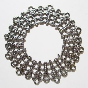 May include: A silver-colored, openwork metal bracelet or decorative element. The circular design features a repeating pattern of ornate, stylized shapes. The piece has a vintage or antique aesthetic, with a detailed, filigree-like construction.