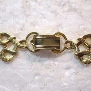 Puede incluir: Cierre de metal dorado con un diseño decorativo. El cierre tiene dos bucles a cada lado y una pieza central rectangular.