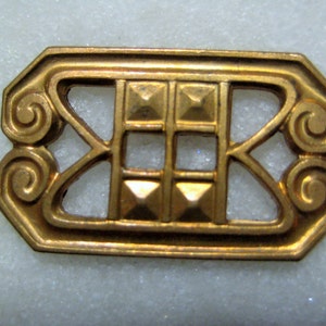 May include: A gold-colored metal decorative piece with a geometric design. The piece has a square shape with a decorative border and a central design of four squares.