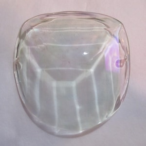 May include: A clear glass paperweight with a faceted design. The paperweight is shaped like a rounded rectangle and has a smooth, polished surface.