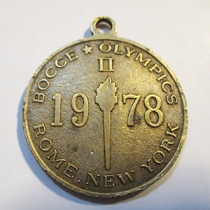 May include: A circular, bronze-colored medal with the text "BOCCE OLYMPICS" and "ROME. NEW YORK" encircling the edges. The year "1978" is prominently displayed, along with a torch symbol. A small loop is at the top for hanging.