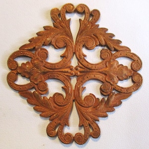 以下が含まれることがあります： 花柄の装飾が施された銅色の金属製の装飾品。十字形に4つのポイントがあり、渦巻きや葉が複雑で細部までこだわったデザインになっています。