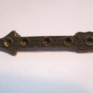 Puede incluir: Un broche de flecha de color dorado con intrincados nudos celtas y cinco acentos circulares.