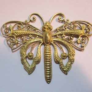 Puede incluir: Un broche de mariposa dorado con intrincados detalles de filigrana. El broche presenta un cuerpo y alas detallados con patrones delicados. Las alas de la mariposa están extendidas, mostrando el diseño detallado. Un accesorio decorativo.