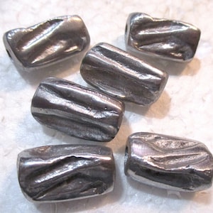Könnte beinhalten: Sechs silberne Metallperlen mit einer strukturierten, gehämmerten Oberfläche. Die Perlen sind zylindrisch geformt und haben eine leicht unregelmäßige Oberfläche.