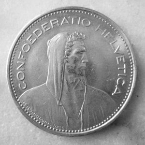Op de afbeelding: Een Zwitserse zilveren munt met de inscriptie "CONFOEDERATIO HELVETICA" en een portret van een persoon met een capuchon.