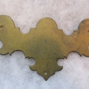 May include: A brass decorative corner bracket with a scalloped edge and three decorative holes. The bracket is a light gold color and has a distressed finish.