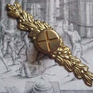 May include: A gold-toned metal decorative piece with a central circular design featuring a cross shape. The piece is adorned with a leafy design on either side.