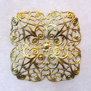 May include: A gold-colored metal filigree design in the shape of a square. The design is intricate and features a repeating pattern of swirls and loops.