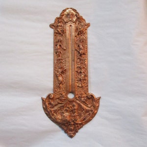 May include: Ornate gold-toned metal thermometer with decorative cherub and floral motifs. The thermometer is mounted on a wall.