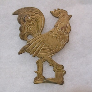 Puede incluir: Una figurita de gallo de metal de color dorado con un diseño detallado. El gallo está de pie con las plumas de la cola extendidas.