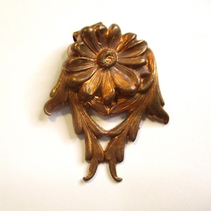 May include: A bronze-colored, ornate decorative element featuring a central flower design with detailed petals. The flower is surrounded by stylized leaves and flourishes, creating a symmetrical, vintage-inspired aesthetic. The piece has a slightly aged appearance.