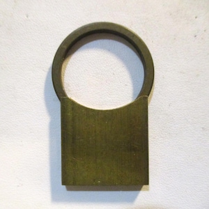 Puede incluir: Un anillo de metal color latón con una base rectangular. El anillo es circular y está unido a la parte superior de la base rectangular. El objeto es probablemente un asa o un elemento decorativo.