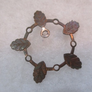 Könnte beinhalten: Ein kupferfarbener Metallanhänger mit einem runden Design mit fünf blattförmigen Ausschnitten. Der Anhänger hat oben eine kleine Schlaufe zum Anbringen einer Kette.