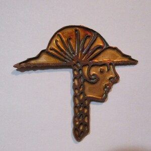 Peut inclure: Une épingle en métal de couleur cuivre avec une image stylisée d'une tête de femme portant un chapeau avec un motif de soleil. Les cheveux de la femme sont tressés et coulent le long de la nuque.
