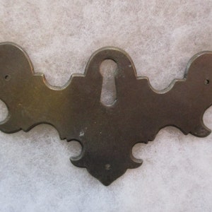 May include: A decorative metal keyhole cover with a ornate design. The cover is made of a dark metal and has a keyhole in the center.