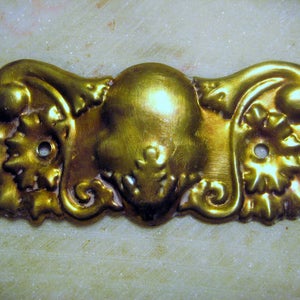 Könnte beinhalten: Ein goldfarbener, verzierter dekorativer Möbelgriff aus Metall. Das rechteckige Stück weist ein zentrales abgerundetes Design mit floralen und Schnörkeldetails an jedem Ende auf. Zwei kleine Löcher sind zur Montage sichtbar.