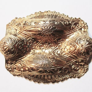 May include: A gold-toned metal brooch with an intricate, embossed design. The brooch has a rounded, oval shape and features a repeating floral pattern.