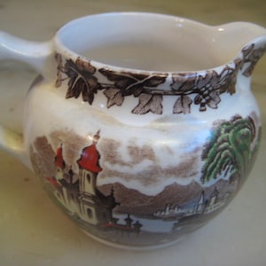 Puede incluir: Una jarra de crema de cerámica blanca con un diseño floral marrón y verde. La jarra presenta una escena de una ciudad con una iglesia y un río.