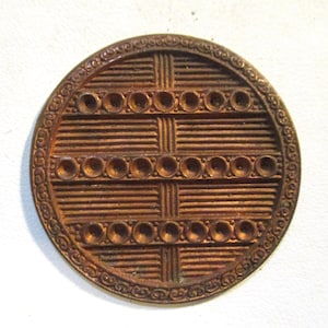 May include: A round, bronze-colored button with a detailed design. The button features a woven pattern in the center, surrounded by a decorative border. Small, circular indentations are evenly spaced throughout the woven section.