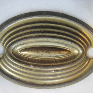 May include: Oval-shaped, gold-toned metal plate with a raised, concentric circle design. The plate has two small holes on either side.